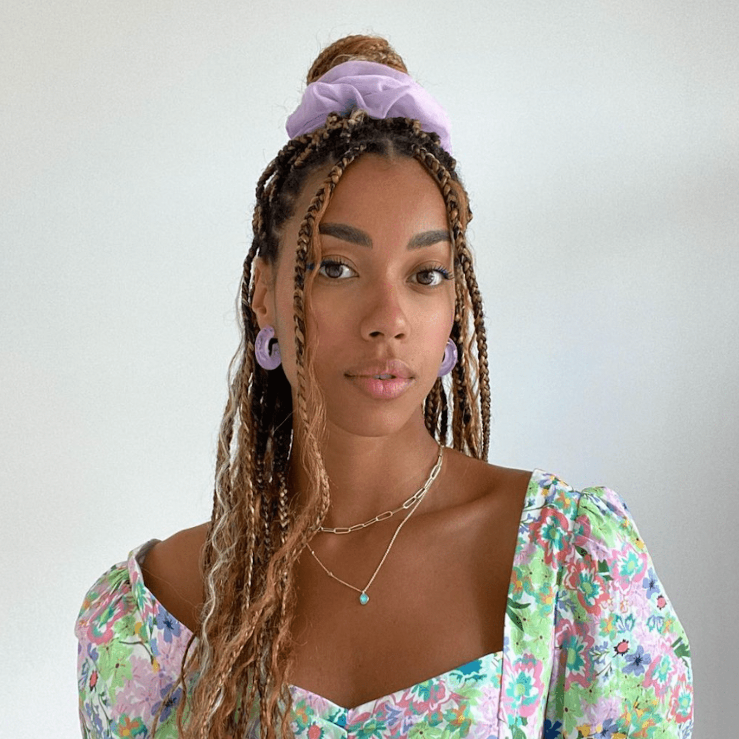 Amaka wears an 80s neon floral dress with lavender hoops and a large lavender scrunchi to hold half her braids into a top knot