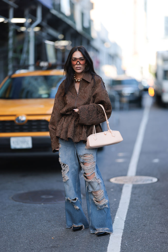 NEW YORK, NEW YORK - SEPTEMBER 09: Olga Sokolenko seen wearing black sunglasses with orange sheer lenses, large gold pearl necklace, Rogov brown pattern embroidered oversized jacket, blue denim destroyed baggy pants / jeans, Alaía beige leather Le Teckel small bag and black shiny leather pumps / heels, during the New York Fashion Week, on September 09, 2024 in New York City.